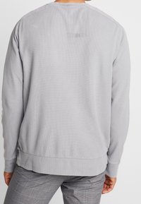 Grauer Pullover mit strukturierter Stoffqualität, gerippten Bündchen und Saum sowie einem Rundhalsausschnitt. Die Rückseite ist glatt und ohne Unterbrechungen.