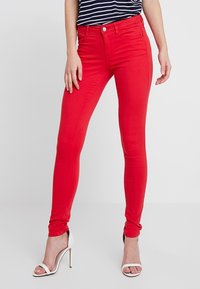 Jeans skinny rouges en denim extensible, présentant une taille mi-haute et un design à cinq poches. Associés à des sandales à talons hauts blanches.