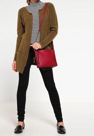 Olijfgroene cardigan met ronde hals, zwart-witte gestreepte coltrui, zwarte legging, en een rode leren schoudertas. Gladde textuur.