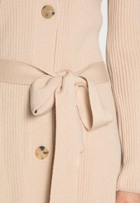 Cardigan in maglia a coste beige con cintura in vita, grande dettaglio con bottone dorato e maniche lunghe, che mette in evidenza la texture e il design.