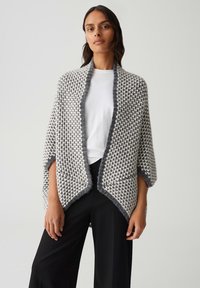 Femme portant un châle en tricot gris et blanc sur une chemise blanche et un pantalon large noir, se tenant devant un fond clair uni.
