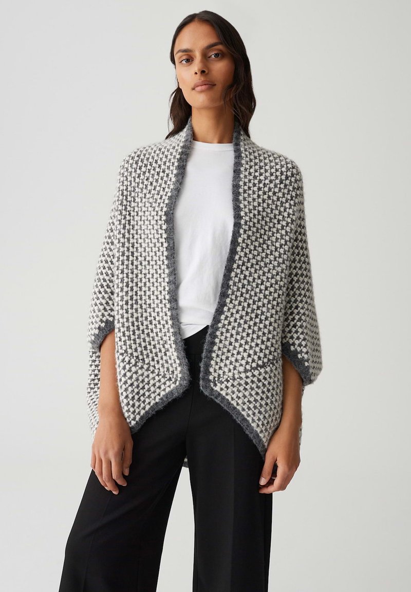 Femme portant un châle en tricot gris et blanc sur une chemise blanche et un pantalon large noir, se tenant devant un fond clair uni.