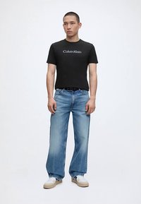 Άνδρας που στέκεται φορώντας μαύρο t-shirt Calvin Klein, ανοιχτό μπλε τζιν με φαρδιά πόδια, λευκά αθλητικά παπούτσια, με απλό φωτεινό φόντο.