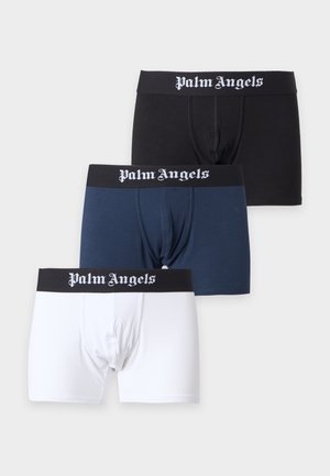 Skjorteløs mand med mørkt hår iført sorte Palm Angels boksershorts står barfodet mod en ensfarvet hvid baggrund.