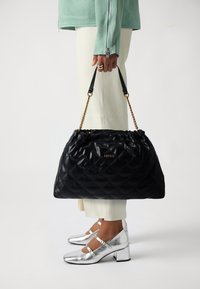 Bolsa de couro acolchoada preta com topo de cordão e detalhes em corrente dourada, usada num ombro, combinada com sapatos de tacão bloco prateados.