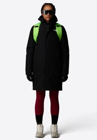 Zwarte lange jas met hoge kraag, neon groene rugzak, zwarte zonnebril, rode legging en beige enkelboots. Geweven stof, gestroomlijnd ontwerp.