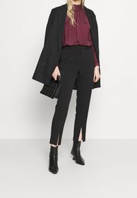Blazer nero, blusa bordeaux e pantaloni neri sartoriali con spacchi laterali. Abbinati a stivaletti neri e una borsa trapuntata.