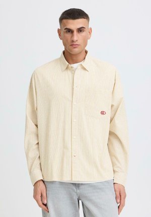 Ung mand iført en beige, stribet skjorte med knapper og et lille rødt "SD"-logo på brystlommen, kombineret med lysegrå jeans.