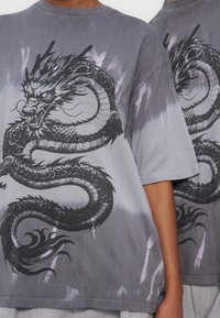 Δύο άτομα φορούν γκρι μπλουζάκια tie-dye με μαύρο, άγριο περιελιγμένο σχέδιο δράκου μπροστά.