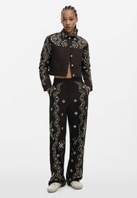 Brązowa kurtka dżinsowa i dopasowane spodnie z białym haftem kwiatowym i falistymi wzorami. Krótki krój, przednie kieszenie i zapięcie na guziki.