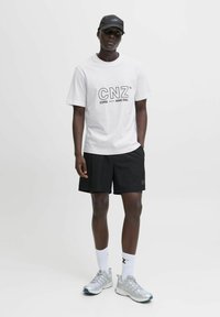 Hvid kortærmet T-shirt med sort "CNZ" grafik, parret med sorte shorts. Grå sportssko og hvide sokker fuldender looket.