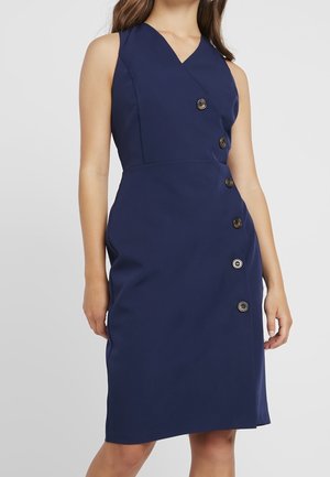 Kobieta ubrana w granatową sukienkę bez rękawów z asymetrycznymi guzikami z przodu i dekoltem w serek.