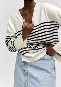 Personne portant un pull blanc à rayures horizontales noires et une jupe en jean bleu clair, ajustant le pull près de la taille.