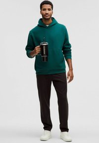 Zelena kapuca s kengursko žepom, crne trenirke i bijele tenisice. Drži crnu bocu za vodu s slamkom i etiketom.