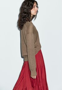 Brązowy dzianinowy sweter o luźnym kroju, zestawiony z zwiewną, plisowaną czerwoną spódnicą. Wyraźnie widoczne są teksturowane włókna oraz swobodna sylwetka.