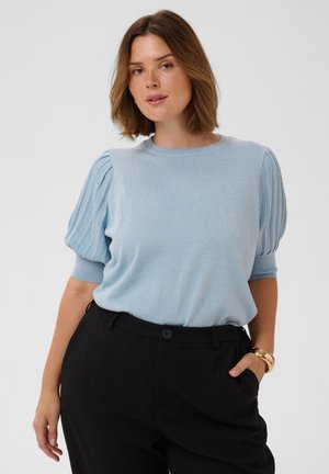 Frau mit schulterlangem braunem Haar, die ein hellblaues kurzärmliges Oberteil mit Puffärmeln und schwarze Hosen trägt, Hand in der Tasche.