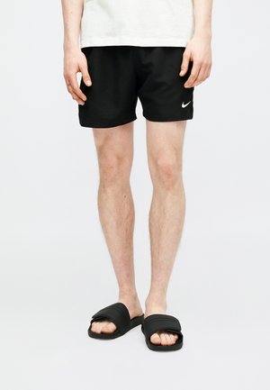 Άτομο που φοράει μαύρα αθλητικά σορτς της Nike και μαύρα σανδάλια, στέκεται μπροστά σε απλό φωτεινό φόντο.