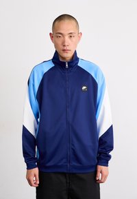 Jaqueta azul-marinho com zíper, com destaques em azul claro e branco, feita de um tecido liso. Apresenta um colarinho alto e um patch de logo na frente.