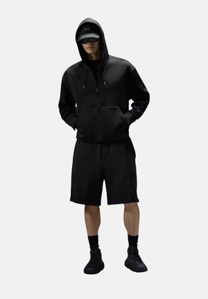 Persona che indossa una felpa nera con cappuccio, pantaloncini neri, scarpe da ginnastica nere, berretto nero e occhiali da sole scuri, in piedi con le mani nelle tasche della felpa.