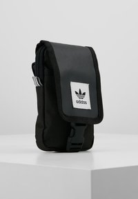 Pochette in tessuto nero con patta pieghevole, dotata di un'etichetta con logo Adidas bianco e una chiusura a clip. Rifinita con dettagli a righe bianche e nere.