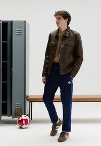 Brun skinnjakke, beige topp, marineblå bukser med blå striper, brune joggesko med hvite striper, fotball, og gråt skap i bakgrunnen.