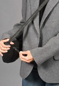 Zwarte leren crossbody tas met een gladde textuur, verstelbare schouderriem en een ritsvak. Model draagt een grijze blazer en denim jeans.