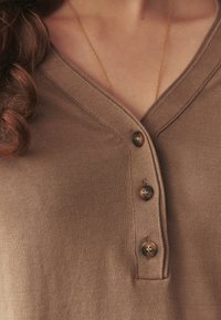 Gros plan d'une personne portant un haut en maille marron boutonné et un fin collier en or, avec des cheveux bouclés reposant sur l'épaule gauche.