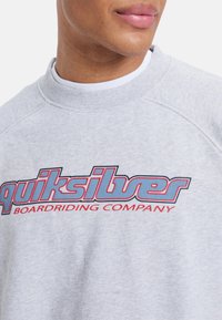 Grå genser med rund hals og raglanerm. "Quiksilver"-logoen i blått med røde detaljer er prominent vist. Myk tekstur.