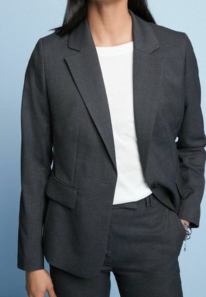 Blazer gris ajustado con un solo botón, solapas de muesca y dos bolsillos laterales. Lleva puesto un camiseta blanca, que presenta una textura suave.