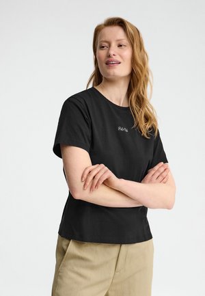 Kvinde med langt rødt hår iført en sort kortærmet skjorte og beige bukser, armene krydset, kigger let til siden mod en ensfarvet baggrund.