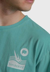 Camiseta de algodón color teal con estampado gráfico plateado que incluye olas, la palabra "Trendsplant" y un icono de elefante circular.