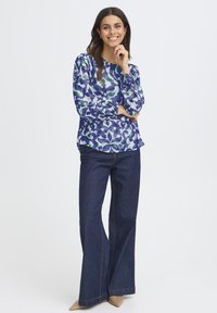 Femme souriante avec de longs cheveux noirs portant un chemisier à motifs bleus, un jean large foncé et des chaussures à bouts pointus beiges, se tenant avec la main près du menton.