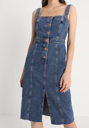 Midi sukienka denim z szerokimi ramiączkami, guzikami z przodu, detalami szwów, paskiem w talii i przednim rozcięciem, noszona przez osobę z ręką spoczywającą przy boku.