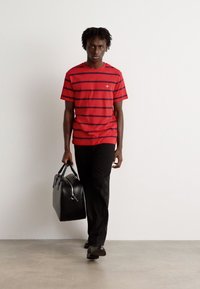 Rød og marineblå stribet kortærmet t-shirt; sorte bukser; sort lædertaske; glat stof; person der går på et gråt gulv mod en ensfarvet baggrund.