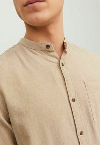 Beige textilní košile s texturou, stojáčkovým límcem, zapínáním na knoflíky a kapsou na prsou. Má hnědé knoflíky a jednoduchý design.