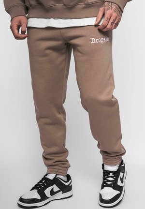 Brown sweatpants with ribbed cuffs; features "Dropsize" logo in white embroidery on the front. Paired with black and white sneakers.