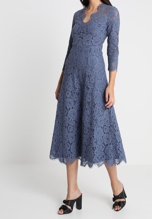 Vrouw draagt een mid-length stoffige blauwe kanten jurk met driekwart mouwen en geschulpte randen, gecombineerd met zwarte sandalen met hak en strikjes.