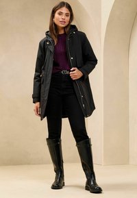 Zwarte waterdichte parka met een trekkoordkap, ongeopende voorkant en gouden hardware, gedragen over een donkerpaars kabelgebreid sweater en zwarte jeans.