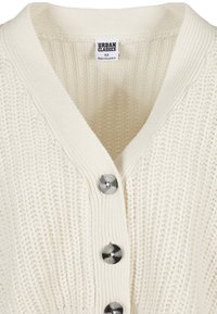 Krämfärgad stickad cardigan med V-ringning, med ribbat mönster och fem rund knappar med metallisk lyster. Urban Classics etikett synlig.