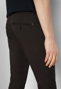 Gros plan sur une personne portant un pantalon noir ajusté avec une poche arrière boutonnée et un petit détail métallique près de la taille.