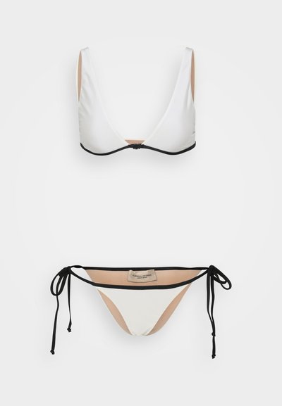 Balta bikini komplekts ar trīsstūra topu ar melnu apdari un regulējamiem aukliņām, kā arī apakšdaļu ar sānu aukliņām, kas arī ir balta ar melniem akcentiem.