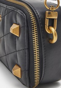 Černá kožená taška s prošívanou texturou, zlatým zipem a zlatými kovovými detaily včetně pyramidových hrotů a klipsového hardwaru po straně.