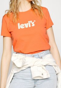 Camiseta de algodón naranja de manga corta con el logotipo de texto blanco "levi's®". Combinada con unos vaqueros de talle alto de color azul claro y una chaqueta crema atada a la cintura.