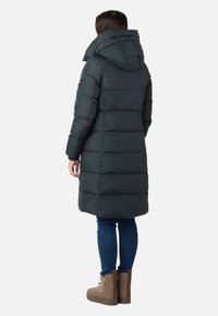 Polstret donkergrå frakke med høj krave og fastsyet hætte. Har horisontale syninger, lange ærmer og går til midt på låret. Bæres med blå jeans og brune støvler.