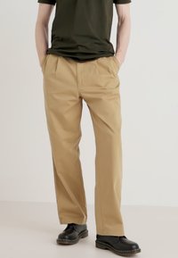 Khaki trousers with a straight cut, front pleats, and side pockets; made from lightweight fabric, paired with black shoes.