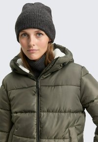 Olijfgroene pufferjack met een ritssluiting aan de voorkant, zwarte accenten en een zachte, witte fleece voering. Gecombineerd met een donkere ribgebreide beanie.