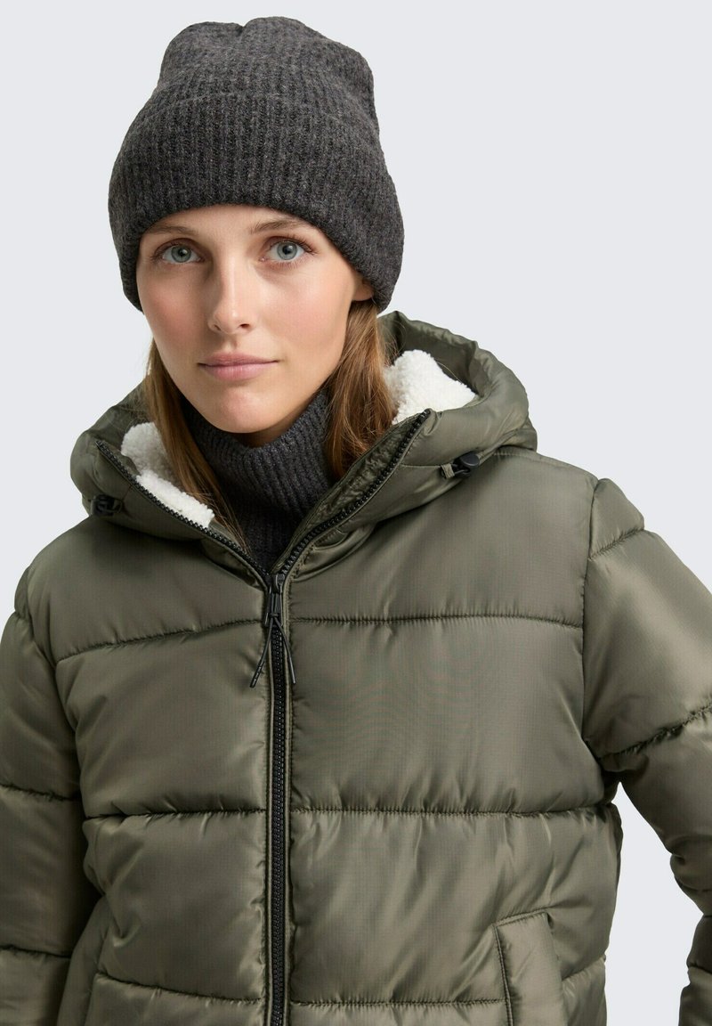 Olijfgroene pufferjack met een ritssluiting aan de voorkant, zwarte accenten en een zachte, witte fleece voering. Gecombineerd met een donkere ribgebreide beanie.