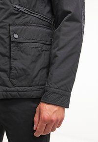 Svart vadderad jacka med en dragkedjeficka och tryckknappdetaljer. Har breda ärmslut med en texturerad finish och en något glansig yta.