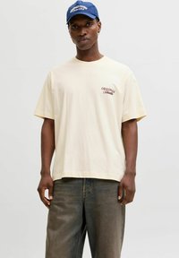 T-shirt κοντομάνικο, απαλό κίτρινο με χαλαρή γραμμή, που διαθέτει ένα μικρό μαρόνο λογότυπο "ORIGINALS" στο αριστερό στήθος. Φοριέται με σκούρα τζιν παντελόνια.