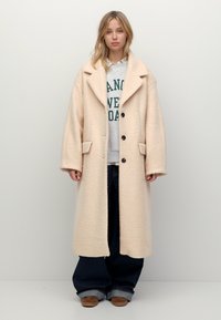 Jasno beżowy, oversize'owy płaszcz wykonany z teksturowanej tkaniny, z dużym kołnierzem, dwiema kieszeniami z przodu i dwoma guzikami. Nosi się go na codzienne ubrania.